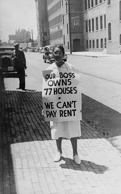 Woman Standing on a Street Holding a Sign Reading “OUR BOSS OWNS 77 HOUSES — WE CAN’T PAY RENT”